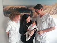 Zwei Personen in medizinischer Kleidung stehen zusammen. Eine dritte Person in der Mitte hält ein Baby in einem weißen Outfit. Im Hintergrund ist ein Bild von einer Landschaft mit Strand und Dünen zu sehen.