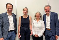 Vier Personen stehen nebeneinander vor einer weißen Wand. Die erste Person trägt einen blauen Blazer und ein weißes Hemd, die zweite Person trägt einen schwarzen Jumpsuit, die dritte Person trägt eine weiße Bluse, und die vierte Person trägt einen blauen Anzug mit einem weißen Hemd.
