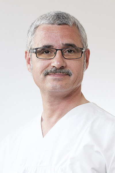 Dr. Johannes Rasbach, Oberarzt und Facharzt für Anästhesiologie, mit grauen Haaren, Brille und weißer medizinischer Kleidung