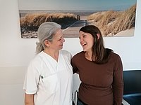 Eine Person in einem weißen Arztkittel steht neben einer Person in einem braunen Oberteil. Im Hintergrund ist ein Bild von einem Strand mit Dünen und einem Holzsteg zu sehen.