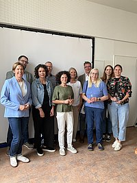 Gruppe von neun Personen steht in einem Raum vor einer weißen Wand