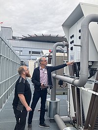 Zwei Personen stehen auf einem Dach neben einer großen Maschine mit Rohren und Steuergeräten. Eine Person zeigt auf ein Bedienfeld.