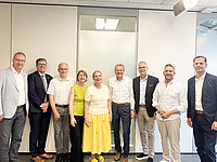 Gruppe von neun Personen steht in einem hellen Raum vor einer weißen Wand und lächelt in die Kamera