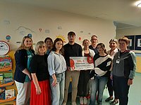 Gruppe von Personen, die vor einer Wand mit Wolken und Sonne stehen. Eine Person hält ein großes Schild mit der Aufschrift '500,- €'.