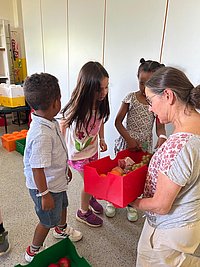 Drei Kinder und eine Frau stehen in einem Raum, die Frau hält eine rote Kiste mit Gegenständen