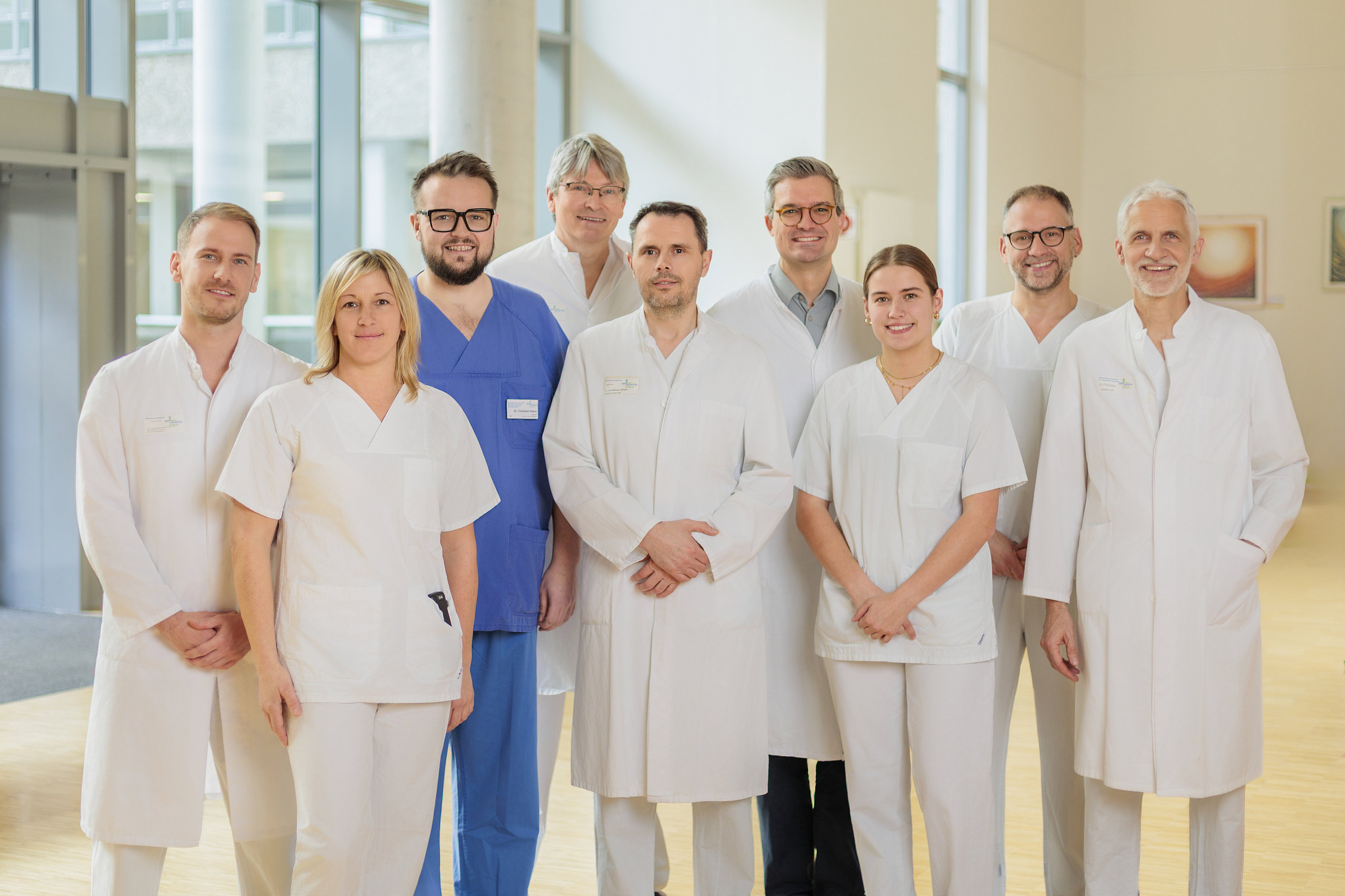 Gruppe von acht medizinischen Fachkräften in weißen Kitteln und Arbeitskleidung in einem hellen Raum