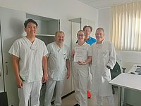 Fünf Personen in weißer Arbeitskleidung stehen in einem hellen Raum mit Schreibtisch und Fenster mit vertikalen Jalousien, eine Person hält eine Urkunde