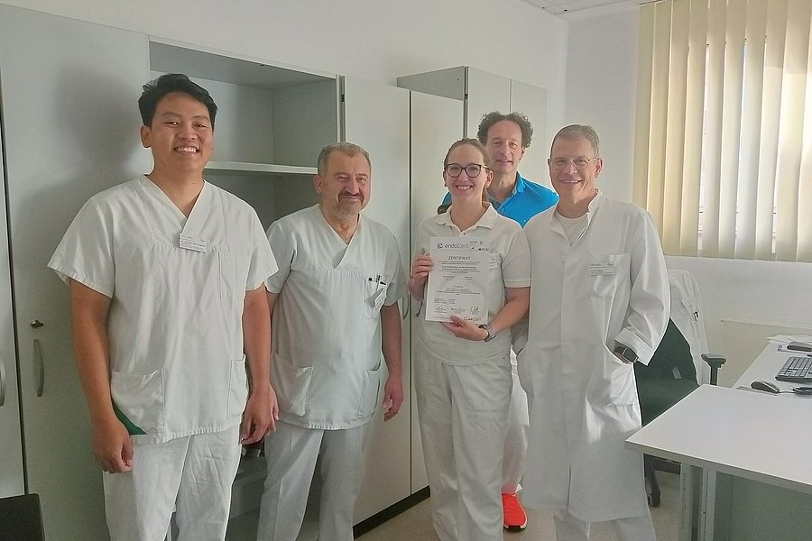 Fünf Personen in weißer Arbeitskleidung stehen in einem hellen Raum mit Schreibtisch und Fenster mit vertikalen Jalousien, eine Person hält eine Urkunde