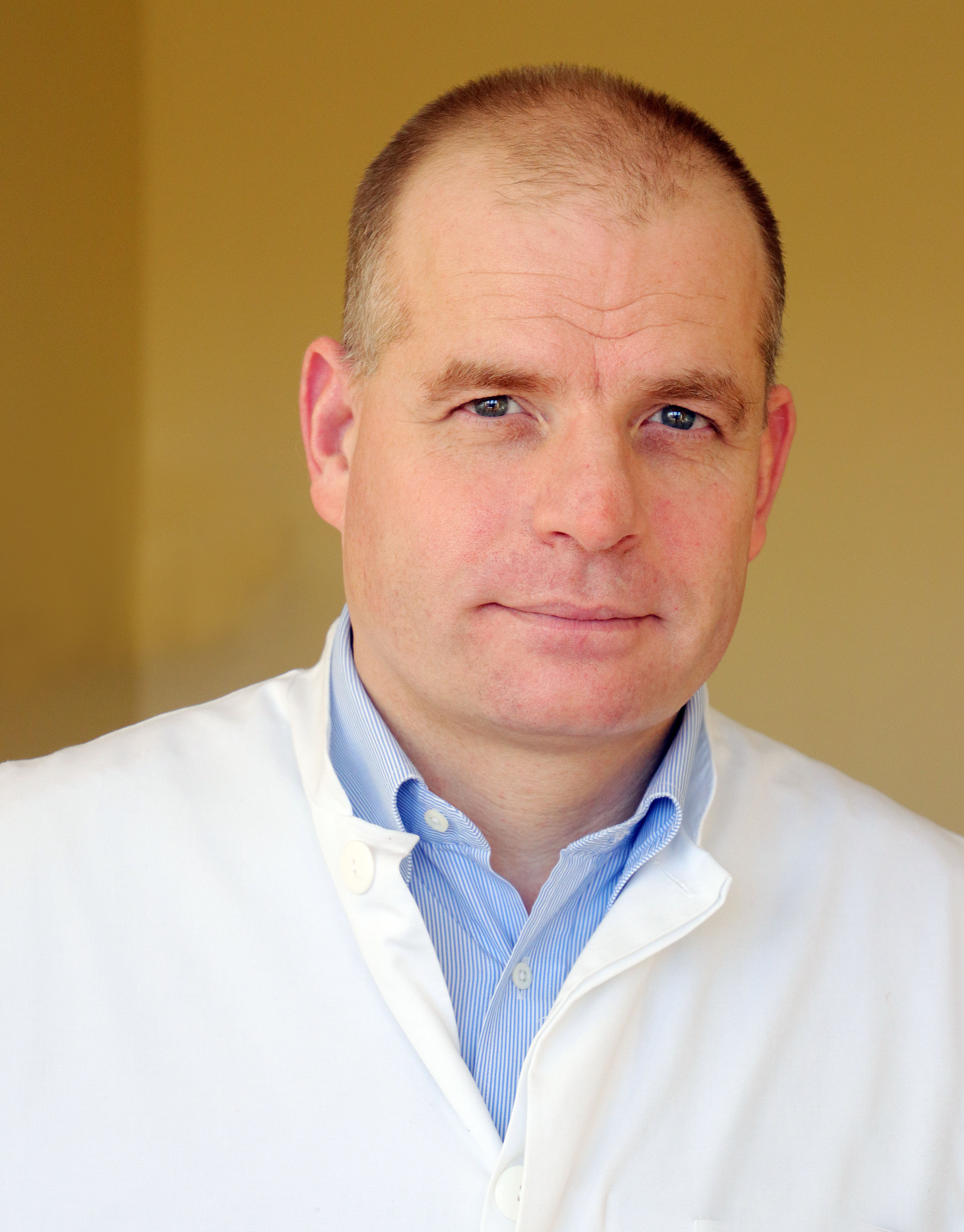 Dr. med. Robert Liszka trägt ein weißes Arztkittel über einem hellblauen Hemd vor beigem Hintergrund.