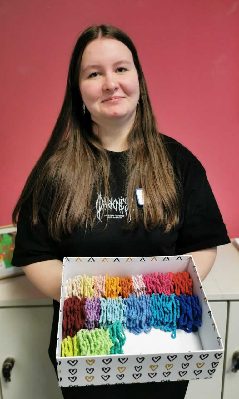 Person mit langen braunen Haaren trägt schwarzes T-Shirt und hält eine Schachtel mit bunten Wollknäueln vor einem roten Hintergrund.