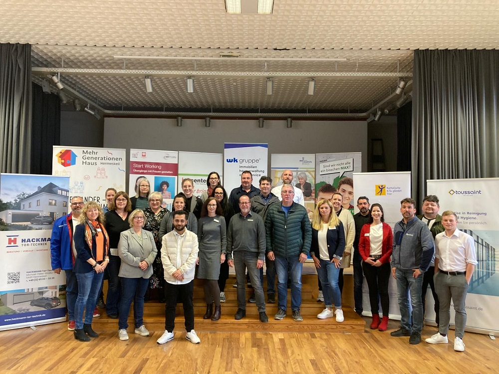 Gruppe von Menschen steht in einem Saal vor mehreren Werbebannern auf einer Bühne mit Holzboden.