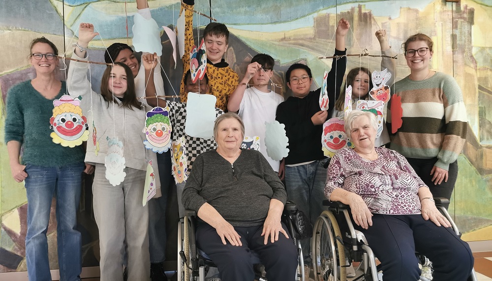 Gruppe von Menschen, darunter zwei Personen im Rollstuhl, halten bunte Clownsmasken an Schnüren hoch, vor einer Wand mit Wandgemälde.