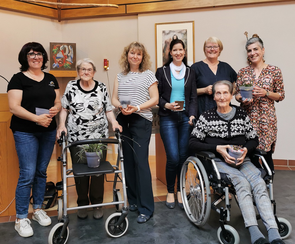 Sechs Frauen in einem Raum, zwei mit Gehhilfen, eine im Rollstuhl, alle halten kleine Schalen, im Hintergrund zwei Bilder an der Wand.