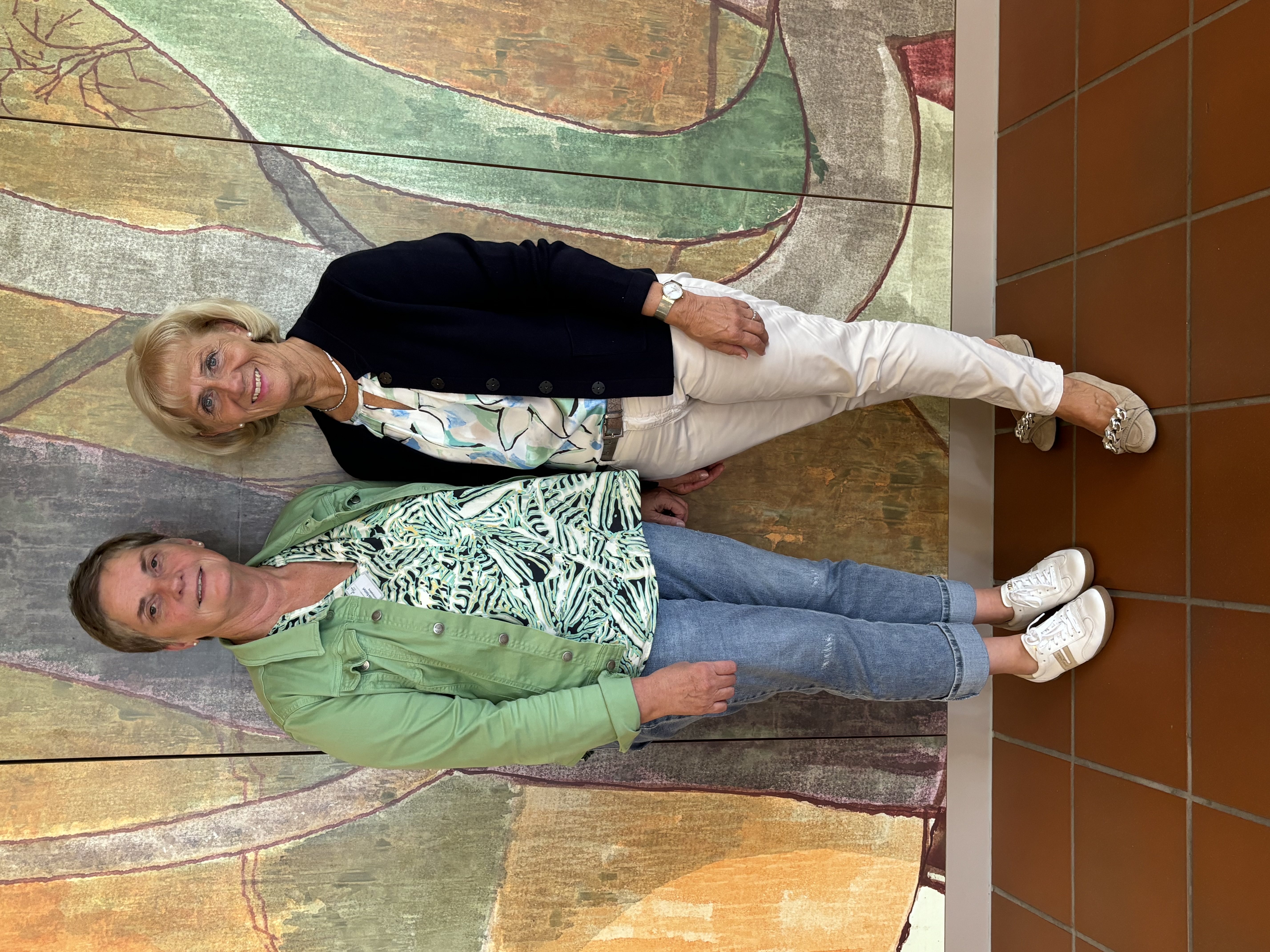 Zwei Frauen stehen vor einer Wand mit abstraktem Wandbild, eine trägt grüne Jacke und Jeans, die andere schwarze Jacke und helle Hose.